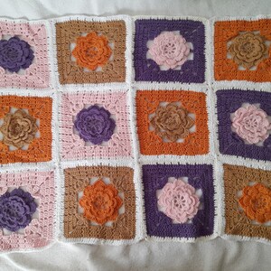 Peut inclure: Une couverture en crochet avec un motif répétitif de fleurs dans des tons de rose, violet, orange et marron. La couverture est composée de carrés individuels cousus ensemble.