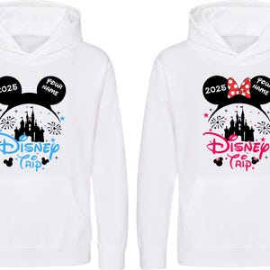 K&ouml;nnte beinhalten: Zwei wei&szlig;e Kapuzenpullover mit einem Disney Trip-Design. Einer hat ein blaues Design, der andere ein rosa Design. Beide zeigen ein Schloss, Feuerwerk und den Text "2025" und "Pour Name" in schwarzen Sprechblasen.