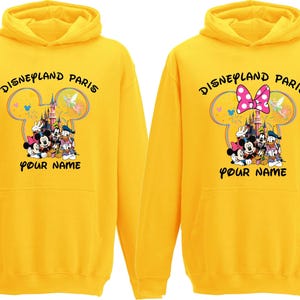 May include: Two yellow hoodies with the text "Disneyland Paris" and a graphic of Disney characters. One hoodie has a pink bow, the other has a Mickey Mouse head design. The text "Your Name" is below the graphic.