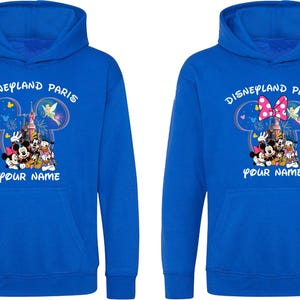May include: Two blue hoodies with the text "Disneyland Paris" above a Mickey Mouse head design. The design includes Disney characters and a castle. The text "YOUR NAME" is below the design. One hoodie has a pink bow.