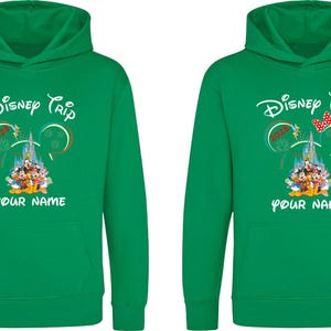 May include: Two green hooded sweatshirts with the text "Disney Trip" and "YOUR NAME" printed in white. The design includes a castle, Mickey Mouse ears, and Disney characters. One sweatshirt has a red bow.
