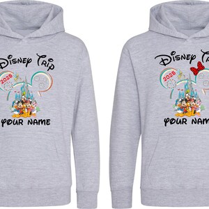 May include: Two light gray hoodies with a Disney Trip 2025 design. The design includes Mickey Mouse ears, a castle, and Disney characters. The text "YOUR NAME" is below the graphic. One hoodie has a red bow.