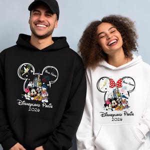 May include: A black hoodie and a white hoodie, each featuring a graphic of Mickey Mouse, Minnie Mouse, Donald Duck, and Goofy in front of a castle inside a Mickey-shaped outline. The text reads Disneyland Paris 2026 and includes a space for a name.