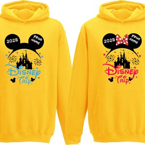 K&ouml;nnte beinhalten: Zwei gelbe Kapuzenpullover mit einem Disney Trip-Design. Einer zeigt eine schwarze Mickey Mouse-Ohren-Silhouette mit dem Text "2025" und "Your Name". Der andere hat eine Minnie Mouse-Schleife und denselben Text. Beide haben eine Schlossgrafik.