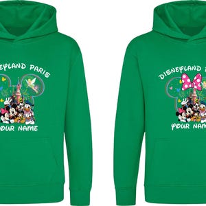 May include: Two green hooded sweatshirts with the text "Disneyland Paris" and a graphic of Mickey Mouse, Minnie Mouse, Donald Duck, and other characters. One has a pink bow. The text "YOUR NAME" is below the graphic.