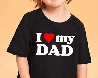 May include: A black t-shirt with the text "I ❤️ my DAD" in white letters and a red heart. The shirt is worn by a child with blonde hair and blue jeans. The background is a solid light orange color.