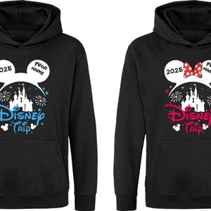 K&ouml;nnte beinhalten: Schwarze Kapuzenpullover mit einem Disney Trip-Design. Einer hat blauen Text und Akzente, der andere rosa. Beide haben eine Schlossgrafik, Mickey Mouse Ohren und das Jahr 2025. Das Design beinhaltet Platz f&uuml;r einen Namen.