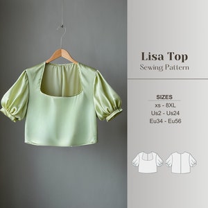 May include: A pale green satin Lisa Top with a square neckline and short, puffed sleeves hangs on a wooden hanger. The sewing pattern includes sizes xs-8XL, Us2-Us24, and Eu34-Eu56. The text "Lisa Top Sewing Pattern" is displayed.