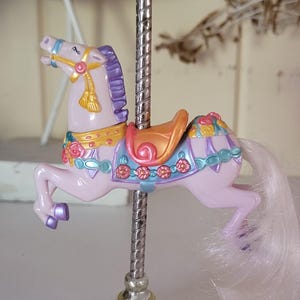 May include: A carousel horse figurine in light purple with a purple mane and tail, featuring a gold saddle, floral accents, and a silver pole. The base is gold-coloured with a banner that reads Morning Glow in script lettering on the front.