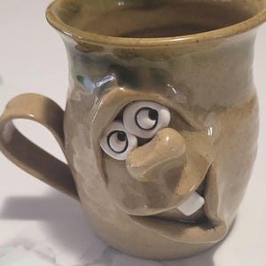 May include: A tan ceramic mug with a sculpted face featuring two white protruding eyes with black pupils, a prominent nose, and a single white tooth visible in the mouth. The mug has a smooth, glossy finish and a curved handle on the left side.