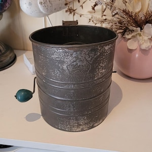 May include: A vintage metal flour sifter with a cylindrical body, a side-mounted crank handle featuring a teal knob, and a stamped logo on the front that reads Kande Patented 7451/26. The metal surface shows signs of wear and oxidation.