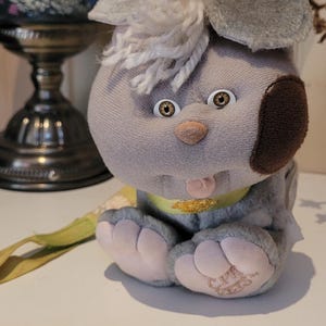 May include: A grey plush toy dog with a large head, white yarn hair, and a dark brown patch over one eye. It features light brown eyes, a small pink nose, a protruding tongue, and a yellow collar with a tag. The text CPK PETS is on its foot.