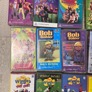 Op de afbeelding: Een verzameling VHS-banden met kinderprogramma's, waaronder Hi-5, Bob de Bouwer en The Wiggles. De banden zijn in een rij gerangschikt, met de titels en logo's duidelijk zichtbaar.