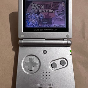Puede incluir: Una consola Game Boy Advance SP plateada, abierta y mostrando el juego "Dragon Ball Z: Buu's Fury". La pantalla muestra personajes y opciones del juego. La consola tiene una cruceta, select, start y botones A/B.