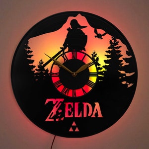 May include: Black wall clock with a silhouette design featuring a mountain scene, trees, and a figure with a sword. The clock face has Roman numerals and the word "ZELDA" in red. The clock is illuminated with red and yellow lights.