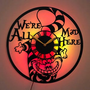May include: A black and white Cheshire Cat wall clock with a red and yellow gradient background. The clock face features Roman numerals and the text "We're All Mad Here".
