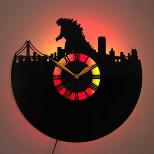 May include: A black vinyl record clock with a silhouette of Godzilla and a cityscape, including a bridge, against a red and orange background. The clock face is illuminated with red and orange lights.
