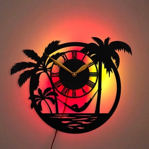 Könnte beinhalten: Eine schwarze Silhouette einer Wanduhr mit Zifferblatt und Palmen. Das Zifferblatt der Uhr hat einen goldfarbenen Stunden- und Minutenzeiger. Die Uhr wird mit einem roten und orangefarbenen Schein beleuchtet.