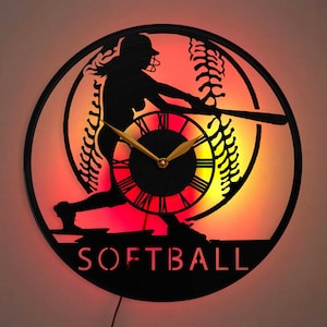 May include: A black and red softball-themed clock with a silhouette of a player swinging a bat. The clock face is white with black Roman numerals and a gold-colored hour and minute hand. The clock has the word "SOFTBALL" written in white letters below the clock face.