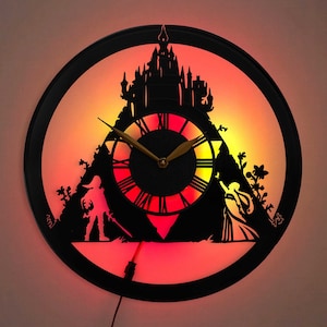 Pode incluir: Um relógio de parede de metal preto com um mostrador de relógio que apresenta algarismos romanos e uma silhueta de um castelo, duas figuras e uma árvore. O mostrador do relógio é iluminado com luzes vermelhas, laranja e amarelas.
