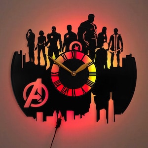 Könnte beinhalten: Eine schwarze Schallplattenuhr mit einer Silhouette der Avengers-Charaktere, darunter Iron Man, Captain America, Thor, Hulk und Black Widow. Das Zifferblatt der Uhr ist ein roter und gelber Kreis mit römischen Ziffern und einem goldfarbenen Zeiger. Die Uhr wird mit roten und gelben LED-Lichtern beleuchtet.
