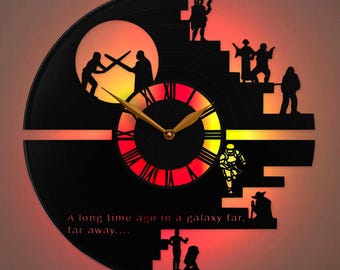 Könnte beinhalten: Eine kreisförmige Wanduhr aus einer schwarzen Schallplatte mit lasergeschnittenen Silhouetten von Figuren und einer Duellszene. Die Mitte zeigt römische Ziffern mit goldenen Zeigern, beleuchtet durch eine rote und gelbe Hintergrundbeleuchtung. Der Text lautet: A long time ago in a galaxy far, far away....