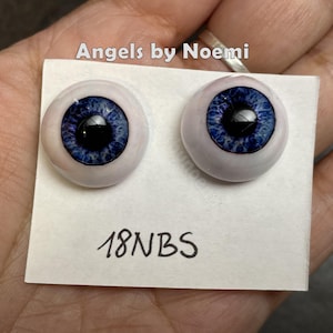 May include: Two round, glass-like doll eyes featuring a deep blue iris with a black pupil and a white sclera. The eyes are mounted on a white rectangular card labelled with the text Angels by Noemi and the alphanumeric code 18NBS.
