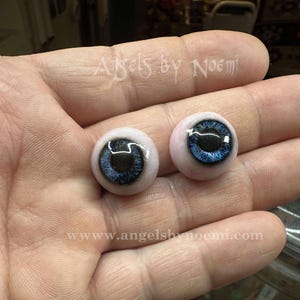 May include: A pair of realistic blue doll eyes with black pupils and a pinkish-white sclera. The eyes are round and appear to be made of glass or a similar material. The text "Angels by Noemi" and website address are visible.