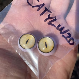 May include: Two round, yellow and black doll eyes with a light purple rim are visible through a clear plastic bag. The bag has the handwritten text "CATYLU20" in purple ink. The eyes have a vertical black slit for the pupil.