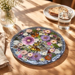 Könnte beinhalten: Ein runder, dekorativer Harztablett mit bunten getrockneten Blumen. Die Blumen umfassen lila Gerbera-Gänseblümchen, rosa Rosen und weiße Gänseblümchen. Ein Glas Wasser, ein Teller mit Brot und eine kleine Vase mit Blumen stehen ebenfalls auf dem Holztisch.