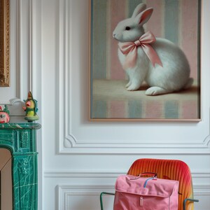 Op de afbeelding: Een wit konijn met een roze strik zit op een roze en blauwe gestreepte achtergrond in een ingelijst schilderij. Het schilderij hangt aan een witte muur in een kamer met een groene marmeren open haard en een roze en oranje gestreepte stoel.