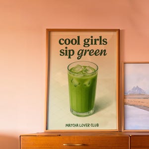 May include: A framed print showing a green matcha drink with ice cubes. The text "cool girls sip green" is above the drink, and "Matcha Lover Club" is below. The print is displayed on a wooden dresser, against a peach-colored wall.