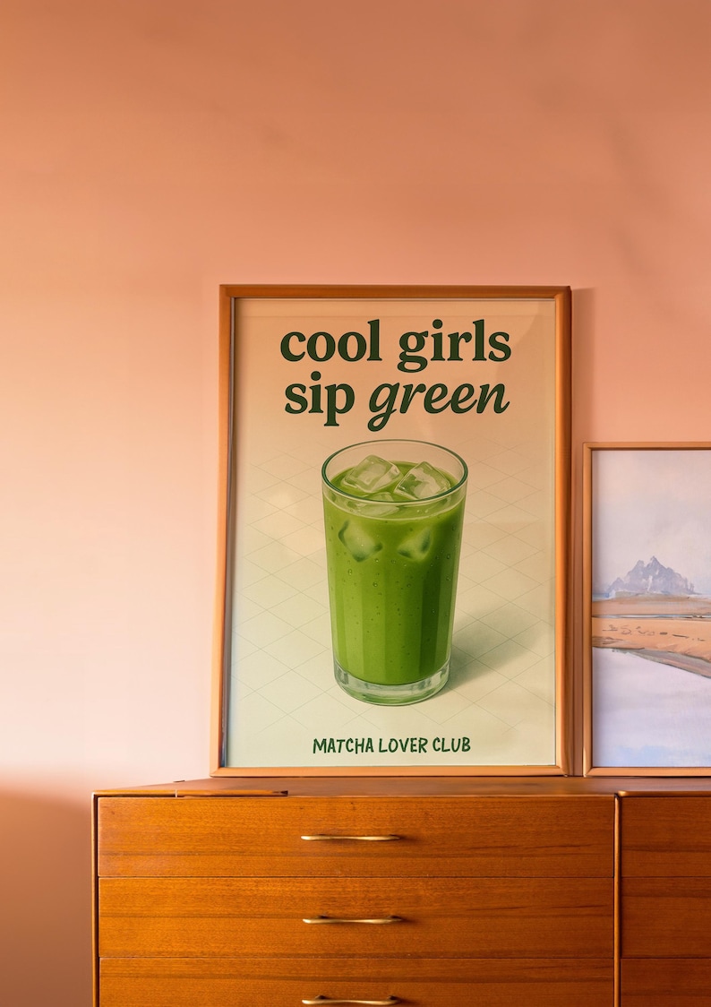 May include: A framed print showing a green matcha drink with ice cubes. The text "cool girls sip green" is above the drink, and "Matcha Lover Club" is below. The print is displayed on a wooden dresser, against a peach-colored wall.