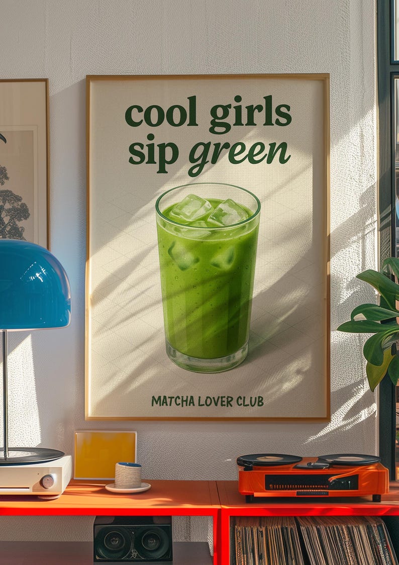 May include: A framed poster with the text "cool girls sip green" and a glass of green matcha drink with ice cubes. The poster is part of a room decor with a record player and other items.