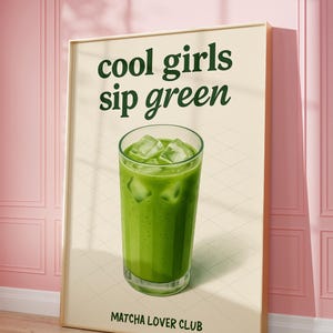May include: A framed poster featuring the words "cool girls sip green" above a glass of green drink with ice cubes. The text "Matcha Lover Club" is at the bottom. The poster is propped against a pink wall.