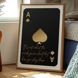 Pode incluir: Uma impress&atilde;o emoldurada de um &aacute;s de paus de baralho com detalhes em folha de ouro. O cart&atilde;o &eacute; preto com um paus dourado e o texto "It's not about the cards you're dealt but how you play them" em letras douradas.