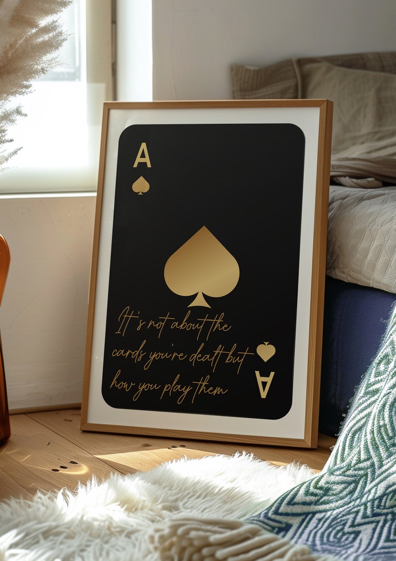 Pode incluir: Uma impress&atilde;o emoldurada de um &aacute;s de paus de baralho com detalhes em folha de ouro. O cart&atilde;o &eacute; preto com um paus dourado e o texto "It's not about the cards you're dealt but how you play them" em letras douradas.