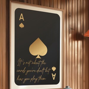 Pode incluir: Um p&ocirc;ster preto e dourado emoldurado com um &aacute;s de paus dourado de baralho e o texto "N&atilde;o se trata das cartas que voc&ecirc; recebe, mas de como voc&ecirc; as joga."