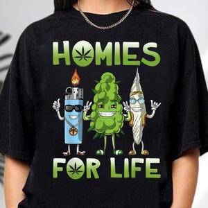 A black t-shirt with a graphic design featuring three anthropomorphic objects: a lighter, a grape, and a surfboard, along with the text "HOMIES FOR LIFE".