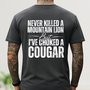 May include: Dark gray t-shirt with white text that reads "NEVER KILLED A MOUNTAIN LION But I'VE CHOKED A COUGAR". The shirt has short sleeves and a crew neck.