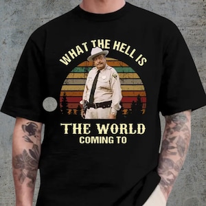 May include: Black t-shirt featuring a vintage-style graphic. The graphic includes a man in a sheriff's uniform, a sunset design, and the text "WHAT THE HELL IS THE WORLD COMING TO".