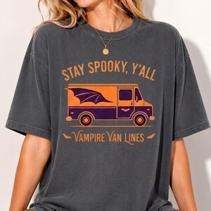 May include: A charcoal grey short-sleeve t-shirt featuring an orange and purple graphic of a van with bat wings. The text reads STAY SPOOKY, Y'ALL and VAMPIRE VAN LINES in orange lettering. The design includes small bat silhouettes and a round gold necklace.