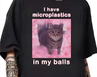 May include: Black t-shirt with the text "I have microplastics in my balls." The shirt features a pink square with a cat image. The cat is gray and brown, set against a pink background with heart shapes.