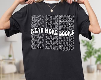 May include: Black t-shirt with the text "READ MORE BOOKS" in a white, wavy font. The text is repeated multiple times down the front of the shirt. The shirt has short sleeves and a crew neck.