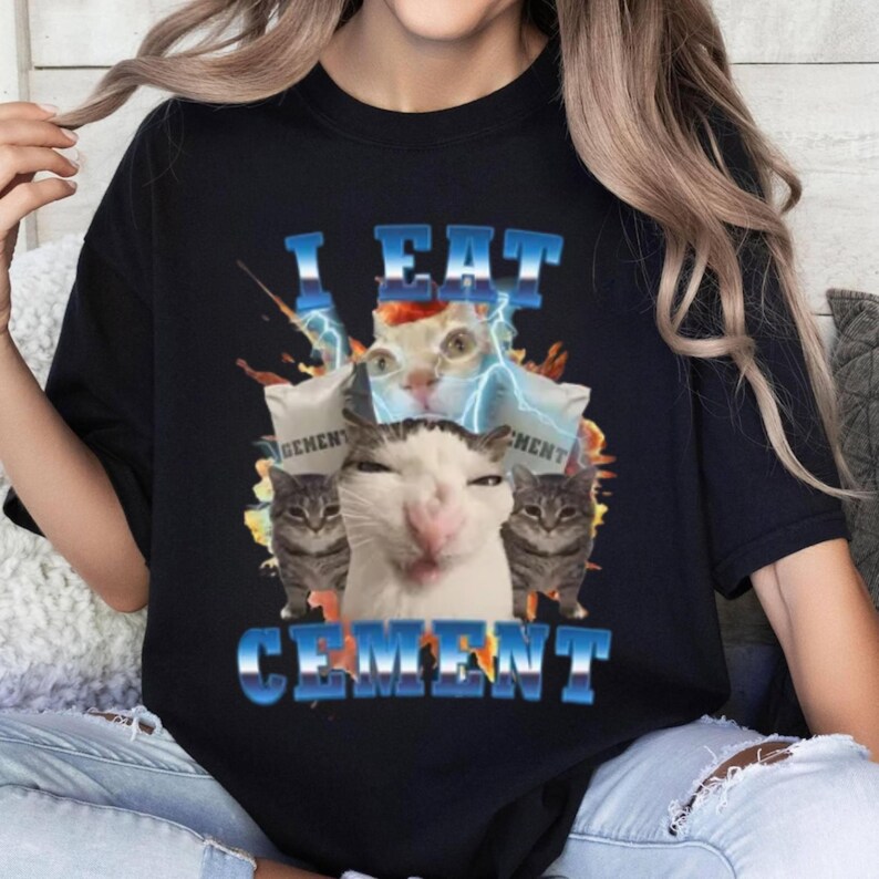 A black t-shirt with a graphic design featuring a cat surrounded by various elements, including a lightning bolt and the text "I EAT CEMENT".