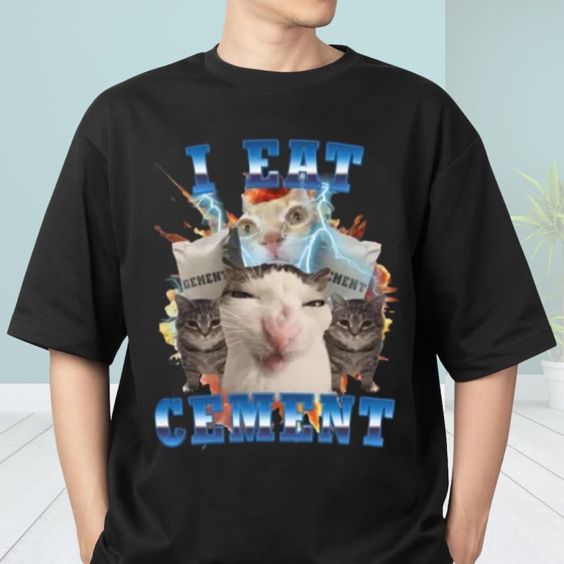 A black t-shirt with a graphic design featuring a cat surrounded by various elements, including a lightning bolt and text that reads "I EAT CEMENT".
