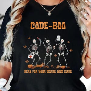 May include: A black t-shirt featuring a graphic of three skeletons dressed as nurses with the orange text CODE-BOO at the top and HERE FOR YOUR SCARE AND CARE at the bottom. The skeletons are surrounded by small orange pumpkins and autumn leaves.
