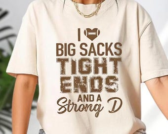 Puede incluir: Camiseta beige con un gráfico marrón. El gráfico incluye un símbolo de corazón de fútbol americano y el texto "I Big Sacks Tight Ends And A Strong D". La camiseta tiene mangas cortas y cuello redondo.
