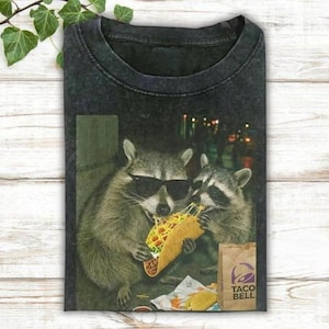 A raccoon wearing sunglasses is eating a taco, with another raccoon sitting next to it.