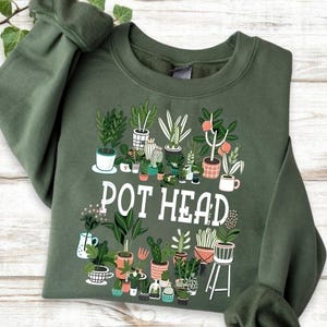 May include: Olive green sweatshirt with a graphic of various potted plants and the words "POT HEAD" in white. The design features cacti, ferns, and other greenery in decorative pots, creating a botanical theme.